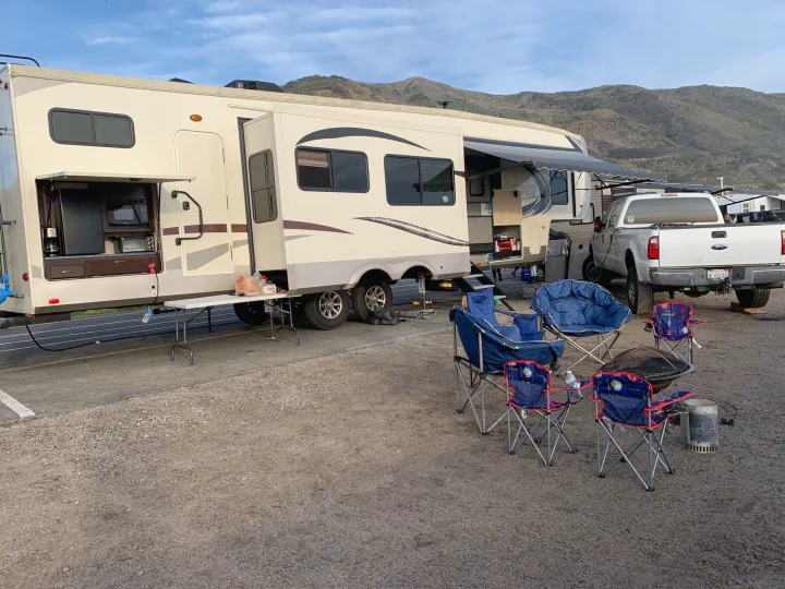 This RV rental setup features a large fifth wheel with an outdoor kitchen, a truck, and camping chairs around a fire pit, backed by mountains.. Palomino Columbus 2018