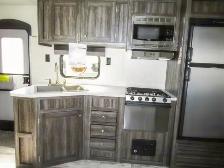 Inside an RV rental: a compact kitchen with wood-grain cabinets, double sink, microwave, stove, and refrigerator.. Openrange 26BHS 2020