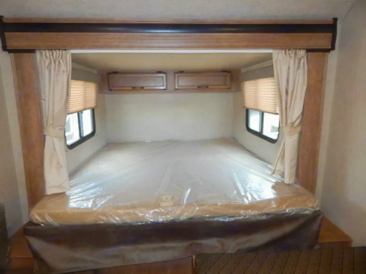 Inside an RV rental: a compact bedroom with a bed protected by plastic, windows with blinds, and wood overhead cabinets.. Starcraft Launch 19BHS 2017