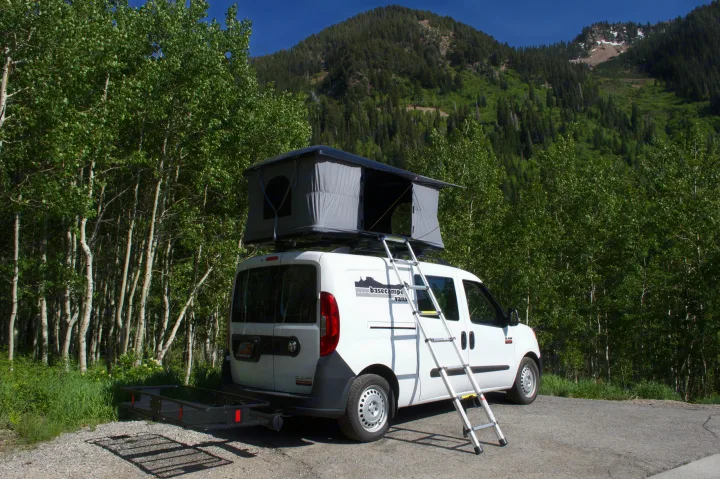White Ram ProMaster City van with rooftop tent, ladder, and cargo carrier in a lush mountain forest. An ideal RV rental for adventure.. Ram Promaster City 2018