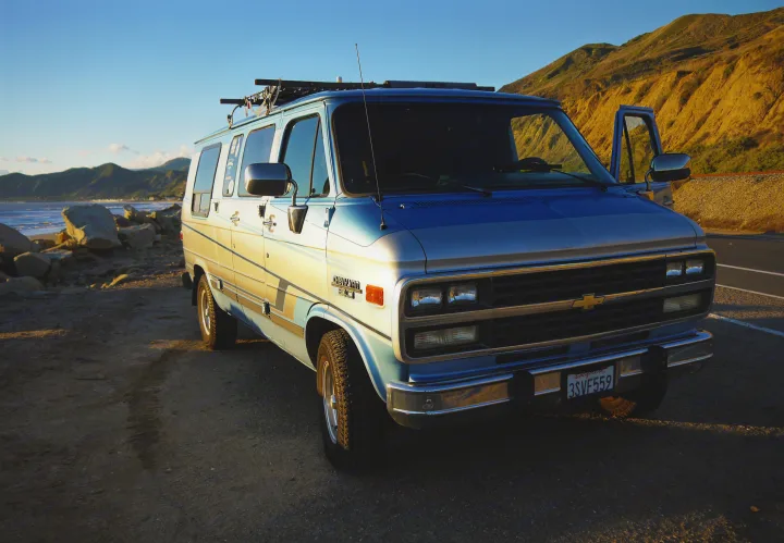 Blue Chevy van parked at a scenic California coast, ideal for an RV rental. Mountains and beach in golden light.. Chevrolet Other 1989