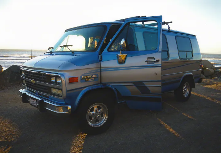 Blue and silver Chevy camper van, likely an RV rental, parked by the ocean with its door open.. Chevrolet Other 1989