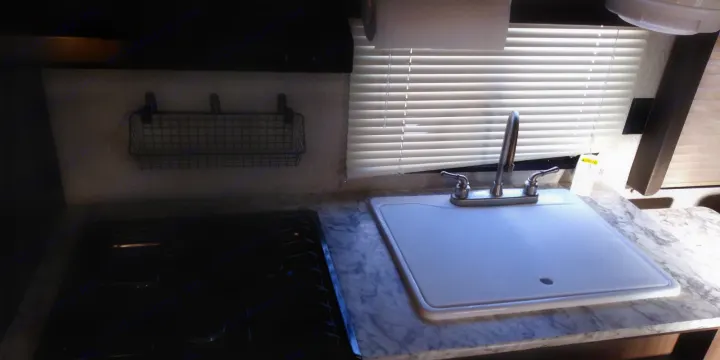 Compact RV rental kitchen with a black stovetop, covered sink, and wall-mounted wire storage basket.. Keystone RV Springdale 2018