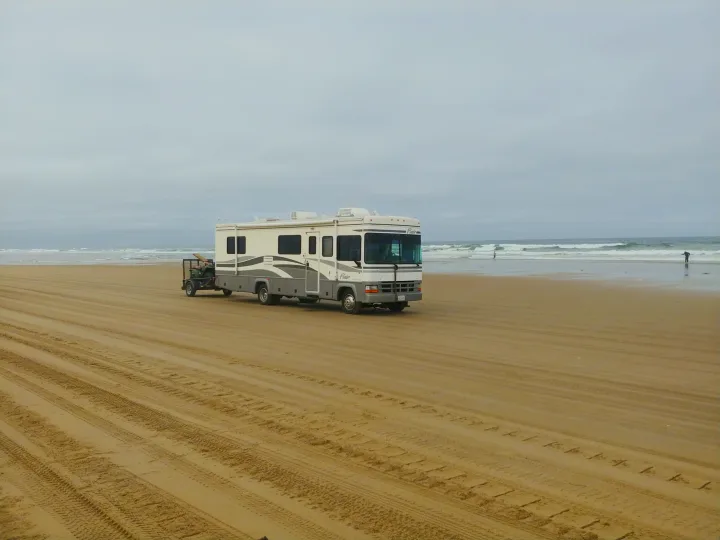 White RV rental on a sandy beach, pulling a trailer with an ATV. Waves crash under an overcast sky.. Fleetwood Flair 2000