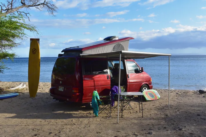 Enjoy beach life! This RV rental, a red camper van with pop-top & awning, is set up on sand by the ocean with a surfboard.. Volkswagen Westfalia 2001