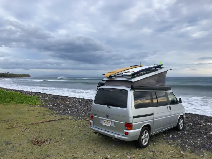 Silver RV rental camper van with surfboards on a rocky beach. Epic waves break under a cloudy sky. Van life & surf travel.. Volkswagen Westfalia 2003
