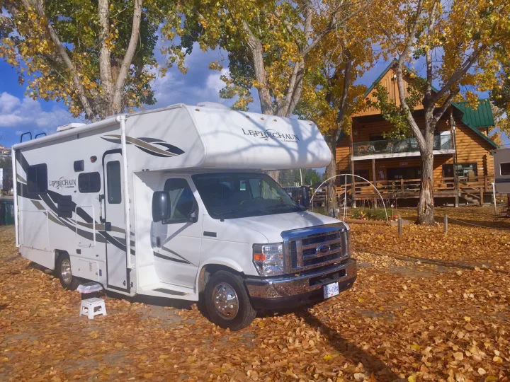 A white Class C RV rental sits amidst a blanket of golden autumn leaves with a rustic log lodge in the background.. Forest River Coachmen Leprechaun 2014