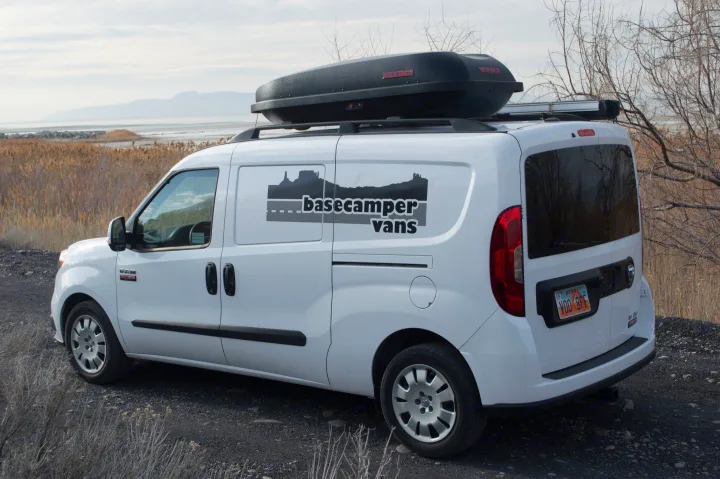 White "basecamper vans" RV rental van with roof box, a Ram ProMaster City, parked by a lake with reeds and mountains.. ram promaster city 2018