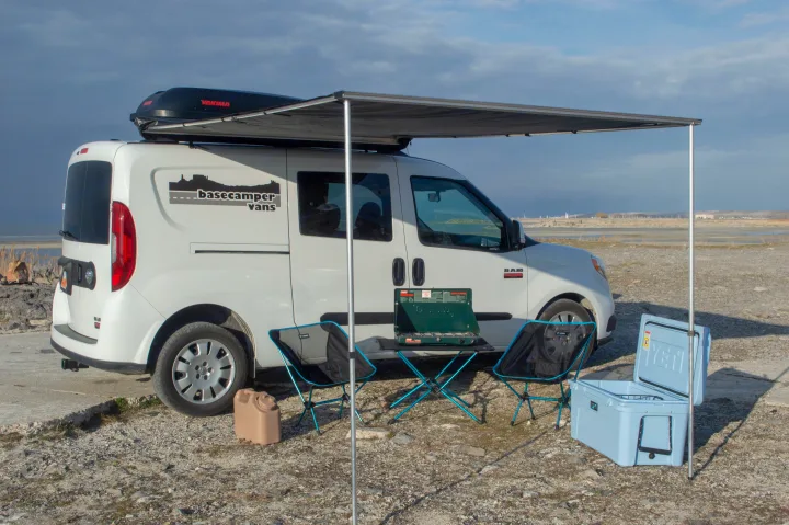 Compact white camper van with an awning extended, two chairs, a stove, and a cooler set up for an RV rental outdoor adventure.. ram promaster city 2018