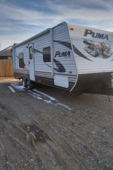 A white Puma travel trailer, ideal for an RV rental, is parked on a gravel lot with patches of snow under a cloudy sky.. Palamino Puma 23FB 2013