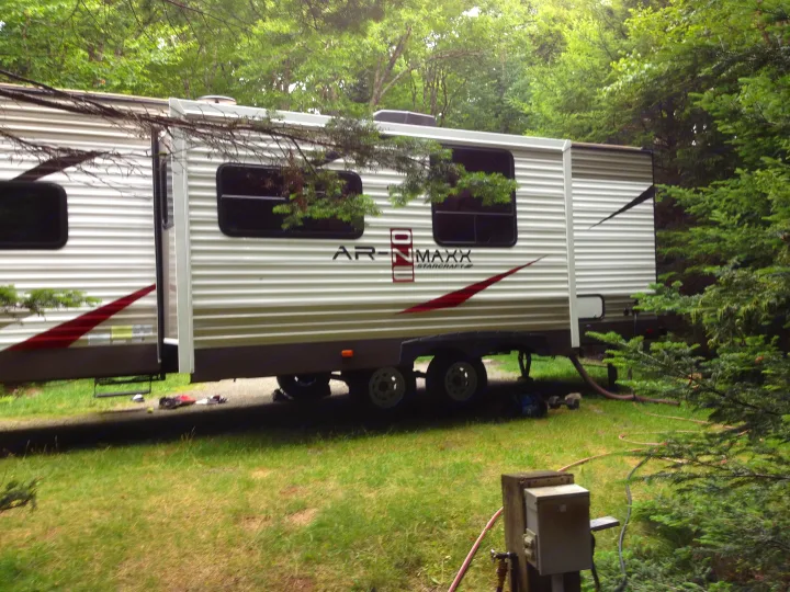 A white RV rental travel trailer with slide-outs extended, parked in a vibrant green, wooded campsite with hookups visible.. Starcraft Ar-One MAXX 2016