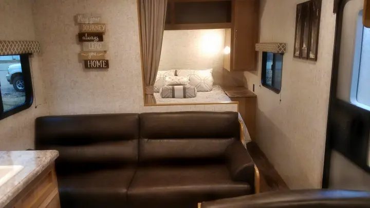 This cozy rv rental features a living space with a brown sofa, a bed alcove with a curtain, and a wall sign reading "HOME".. Braxton Creek 26BH 2019