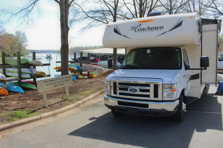 White Coachmen Freelander RV rental parked by a lake, with stacked kayaks, a dock, and covered boat slips in the background.. Coachmen Freelander 2019