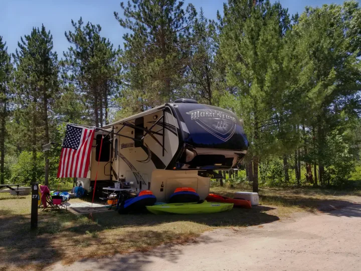 Rv rental: A large Momentum fifth-wheel RV at a wooded campsite (site 27), with an American flag, kayaks, and camping gear.. Grand Design Momentum 385 TH 2015