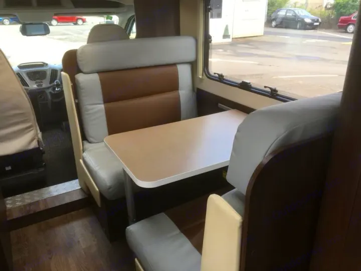 View inside an RV rental: brown & gray dinette with table, leading to the driver's cockpit. Window view on right.. Other Transit Motorhome 2018