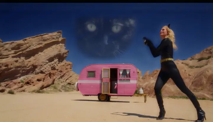 A woman in a catsuit poses with a pink RV rental in a desert with rock formations, a giant cat face in the sky.. Pleasurecraft Canned Ham 1956