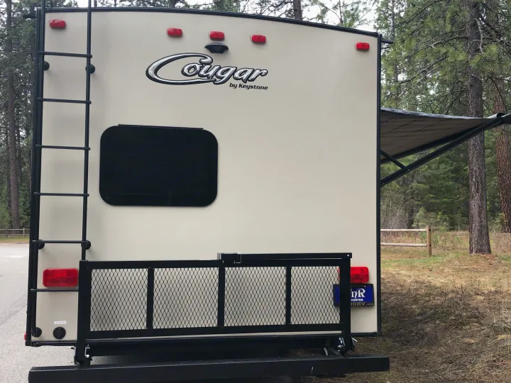 Rear view of a Cougar RV rental with ladder, cargo rack, and awning extended, parked in a wooded camp spot.. Keystone Cougar 2018