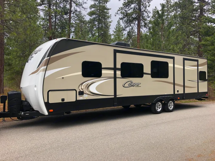 This spacious Cougar travel trailer, ideal for an RV rental, is parked on a paved road surrounded by tall pine trees.. Keystone Cougar 2018