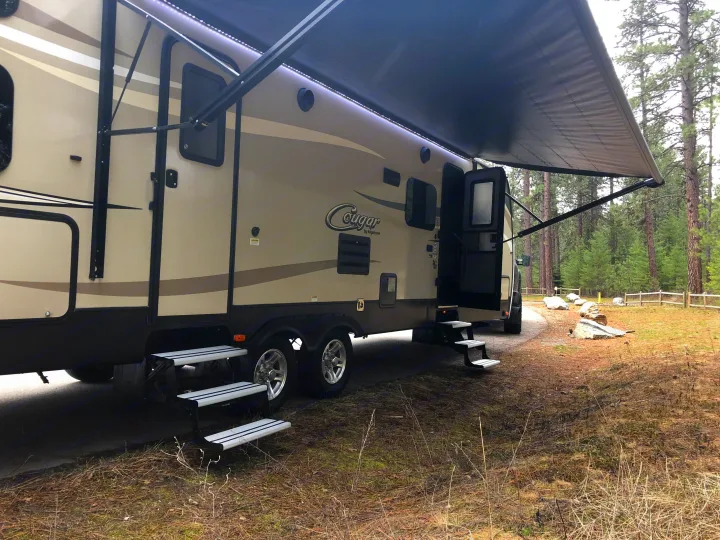 RV rental: A tan and black travel trailer set up in a quiet, wooded campsite with its awning extended and steps down.. Keystone Cougar 2018