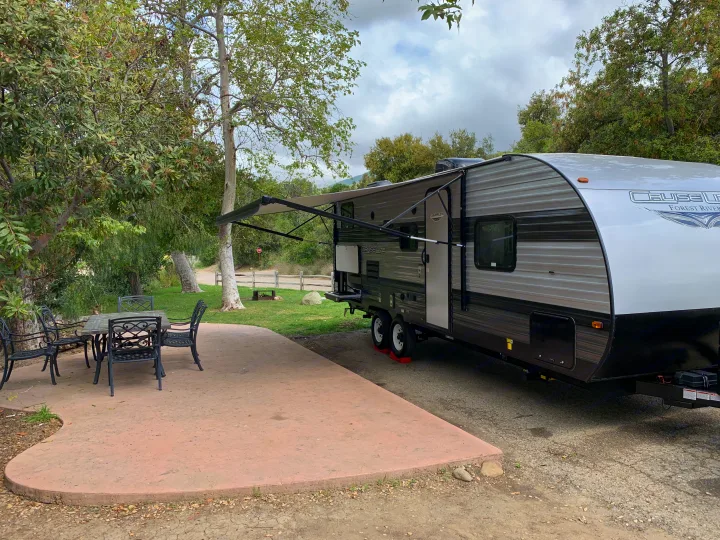 Relaxing RV rental campsite. Features travel trailer with awning, patio, and outdoor seating amidst lush trees.. Forest River Cruise Lite 2020