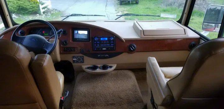 RV rental interior: Driver's seat view of a tan dashboard with wood trim, Ford steering wheel, Jensen stereo, and camera display.. Winnebago Vista 2013