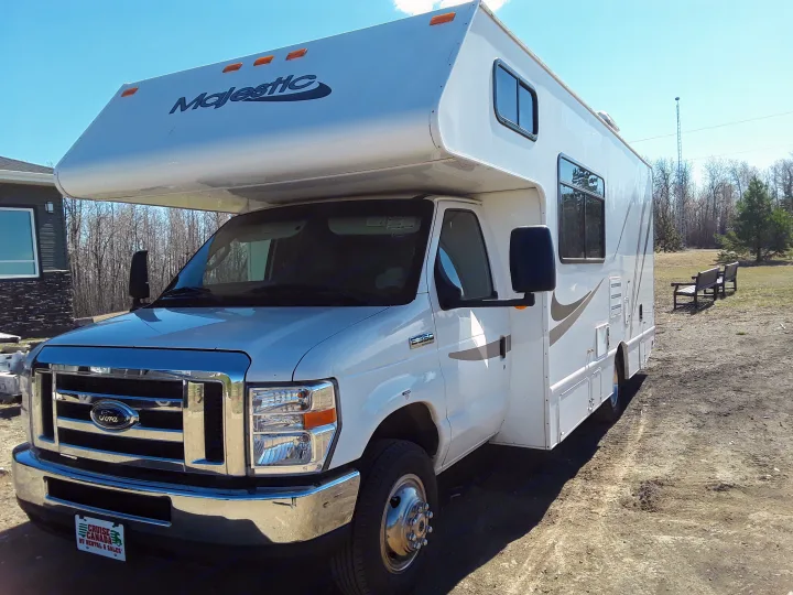 Majestic Class C Ford E-350 RV rental, white and parked on a sunny dirt lot, with trees and benches in the distance.. Thor Motor Coach Four Winds Majestic 2013