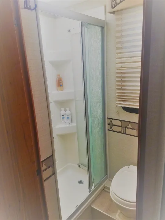 This RV rental bathroom features a compact shower with glass doors, shelves holding toiletries, a toilet, and a window with blinds.. Jayco Melbourne 2017