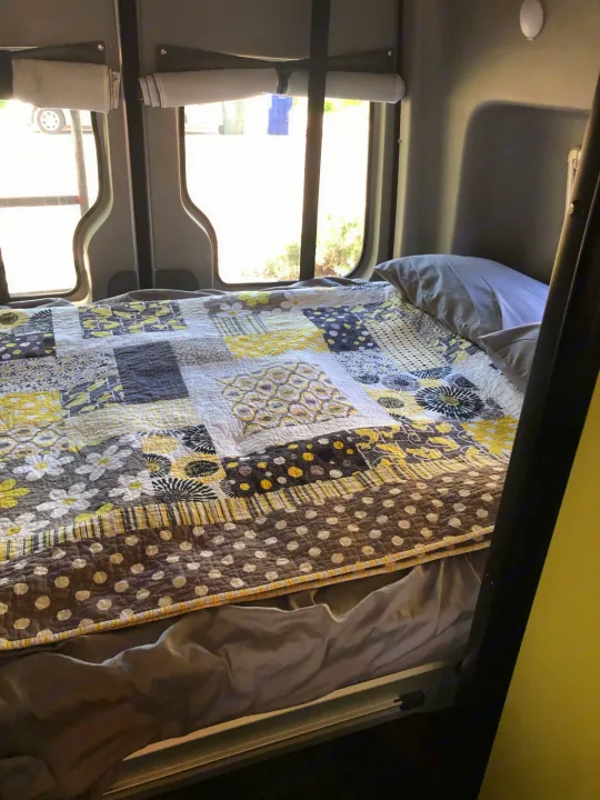 Inside an RV rental: a bright yellow & gray patterned quilt on a cozy bed, with windows and shades visible.. Winnebago Mercedes Sprinter 2019