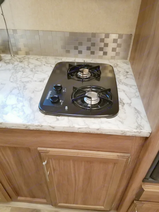 Compact RV rental kitchen with black 2-burner stovetop, marble-look counter, & metallic backsplash.. Jayco Jay Feather 2018