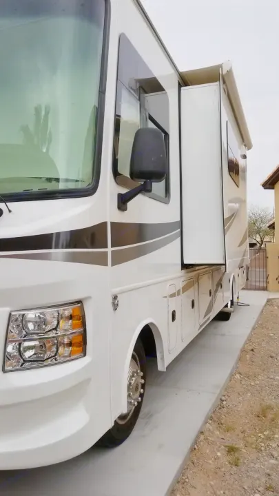 A white motorhome with black stripes and a side slide-out, ready for RV rental or travel, parked next to a wall. Storage bays visible.. Jayco Alante 2018