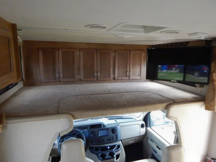 Inside an RV rental, featuring a large overhead bunk, wooden cabinets, TV, and the driver's cockpit.. Gulf Stream Conquest 2016