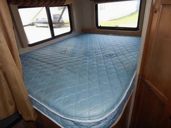 Inside an RV rental: a cozy bedroom with a light blue quilted mattress, framed by windows showing outdoor views.. Gulf Stream Conquest 2016