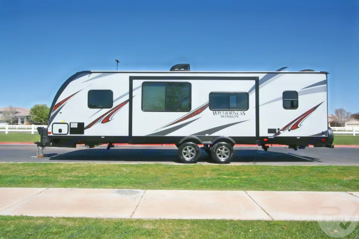A sleek gray and white 'Wilderness Ultralite' travel trailer, perfect for an RV rental, parked on asphalt under a clear blue sky.. Heartland Wilderness 2020