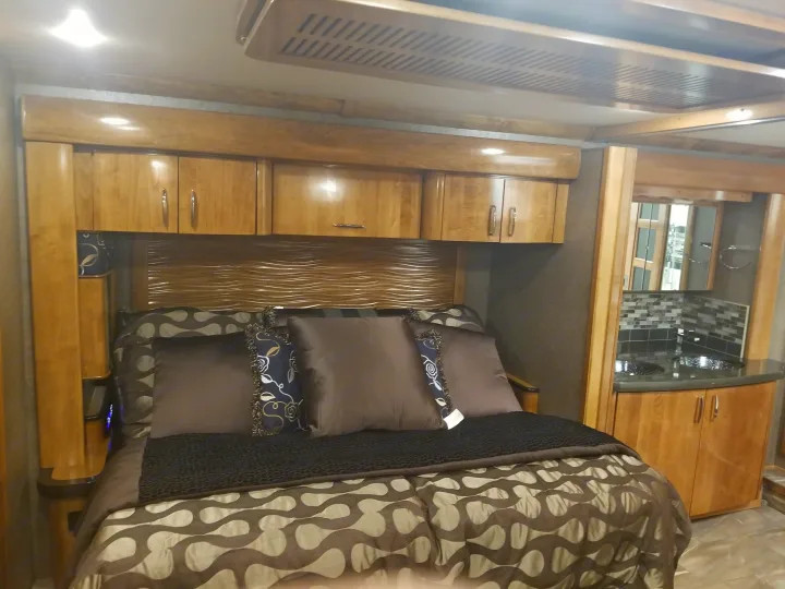 Cozy RV rental bedroom featuring a large bed with patterned bedding, wavy headboard, overhead cabinets, and a compact vanity.. Fleetwood American Eagle 2019