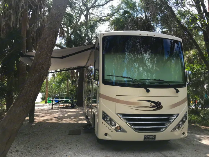 A cream-colored RV rental with an extended awning is parked on a dirt campsite surrounded by trees. A picnic table is seen under the awning.. Thor Hurricane 2018