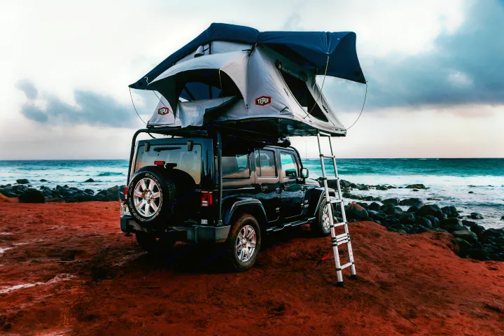 This adventure-ready Jeep Wrangler with a rooftop tent, available for RV rental, is parked on a vibrant red beach near the ocean.. Jeep Wrangler Unlimited 2016