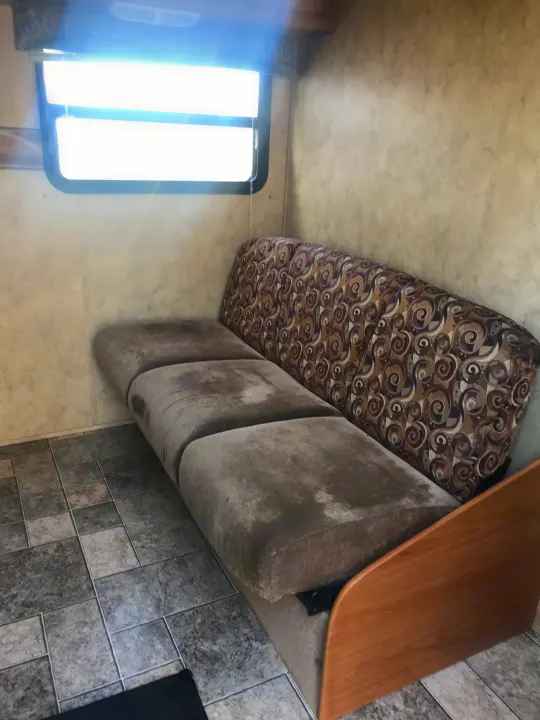 RV rental interior with a well-used sofa, patterned backrest, and bright window. Tiled floor visible.. Keystone Hideout 2009