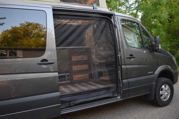 Explore this RV rental's cozy interior: a grey van with its side door open, featuring custom cabinets, microwave, and a bug screen.. Mercedes-Benz Sprinter 2016