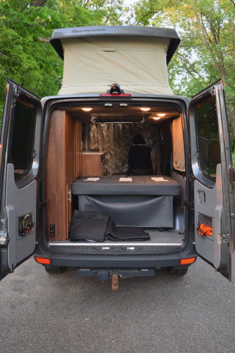 Experience this custom van RV rental! Rear view reveals open doors, pop-top roof, bed, and warm wood interior.. Mercedes-Benz Sprinter 2016