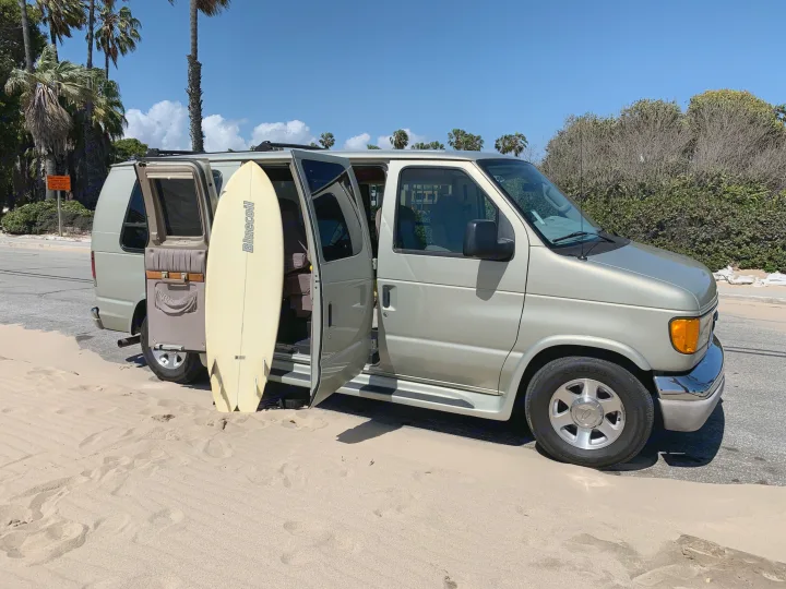 A pale gold RV rental van with open doors and a surfboard stands on a sandy beach next to a road, under palm trees.. Ford Econoline 150 2004