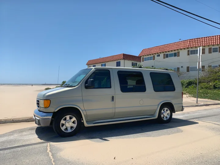 This RV rental van is perfectly parked on a sandy road, ready for a sunny beach day by the ocean and homes.. Ford Econoline 150 2004