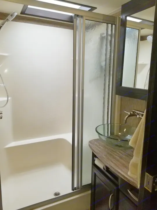 Bright RV rental bathroom with white shower/tub and sliding glass door next to a modern vanity, glass sink, and mirror.. Grand Design Momentum 385 TH 2015