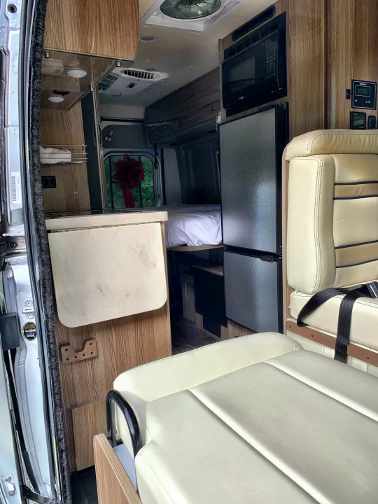 This RV rental interior showcases a compact kitchen with microwave, fridge, and comfortable seating and bed areas.. Winnebago Travato 2019