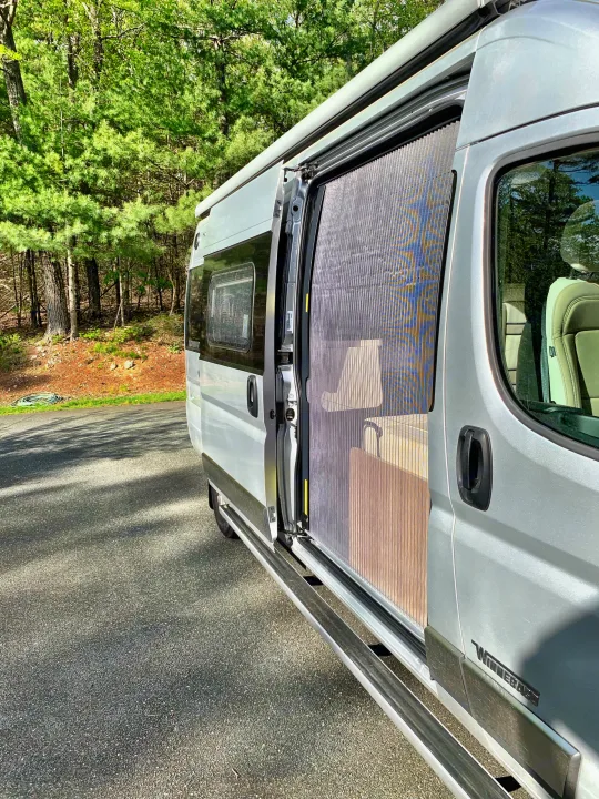 Silver RV rental camper van with open side door and pleated screen, parked on an asphalt lot beside a green forest.. Winnebago Travato 2019
