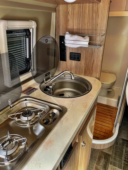 Inside an RV rental: functional kitchen area with a stove & sink, plus a compact wet bath including a toilet and shower.. Winnebago Travato 2019