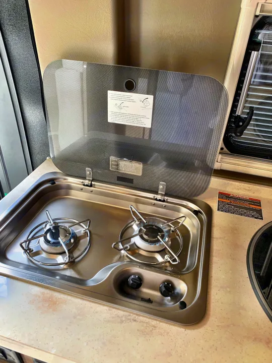 RV rental kitchen featuring a two-burner gas cooktop with an open safety lid, showing usage instructions and a lit burner.. Winnebago Travato 2019
