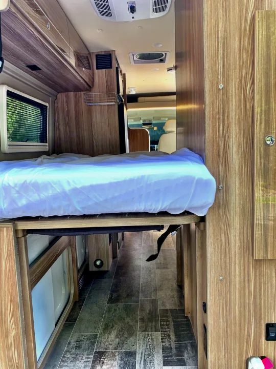 This RV rental offers a practical interior with a raised bed, wood walls, and a clear aisle leading to the front cabin.. Winnebago Travato 2019