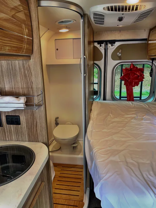Inside this RV rental: a cozy bed, compact kitchen with sink, and a wet bath with toilet. Notice the red bow!. Winnebago Travato 2019
