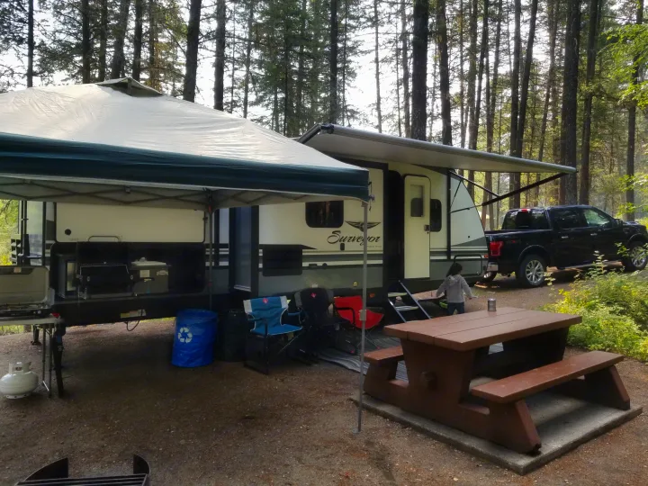 An RV rental parked at a lush, treed campsite with a large canopy, outdoor kitchen, chairs, and a picnic table for comfortable camping.. Forest River Surveyor 2019