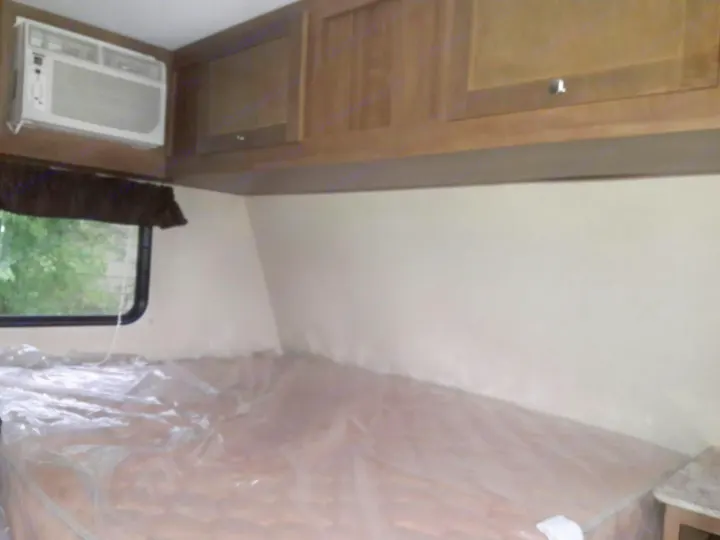 Inside an RV rental: bedroom with a plastic-covered bed, AC unit, window, and wooden cabinets.. Keystone ASPEN TRAIL 1700 2016